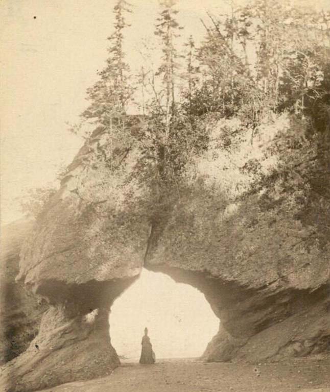 Photographie noir et blanc montrant une arche rocheuse sous laquelle se tient une femme en robe longue sur la plage
