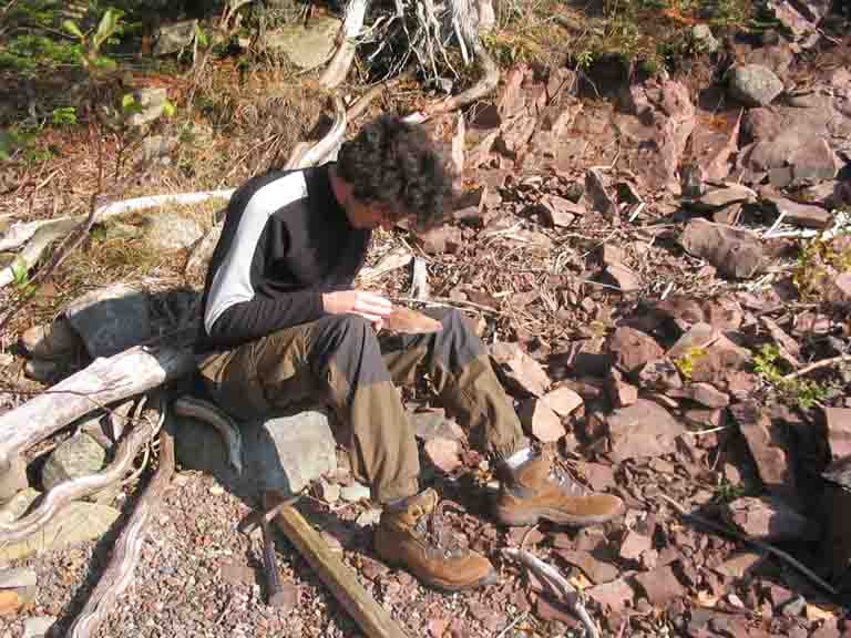Colour photograph of man sitting on rocky shore with rock in his hand