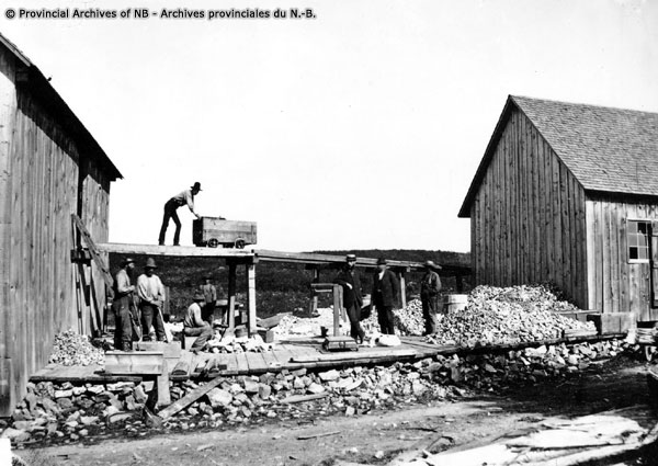 Black and white photograph of rock mine, buildings, sheds, rocks and man on roof of shed