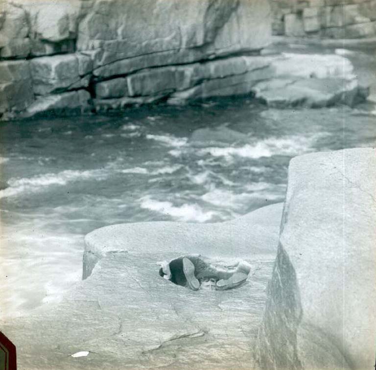 Colour image of river and rocky shoreline with man in a hole with head and feet sticking up