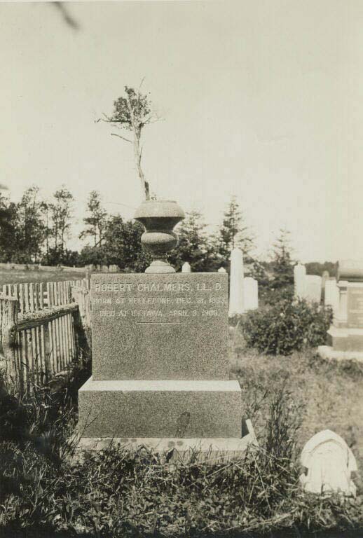 Photographie noir et blanc d’une pierre tombale carrée surmontée d’une urne en pierre dans un cimetière