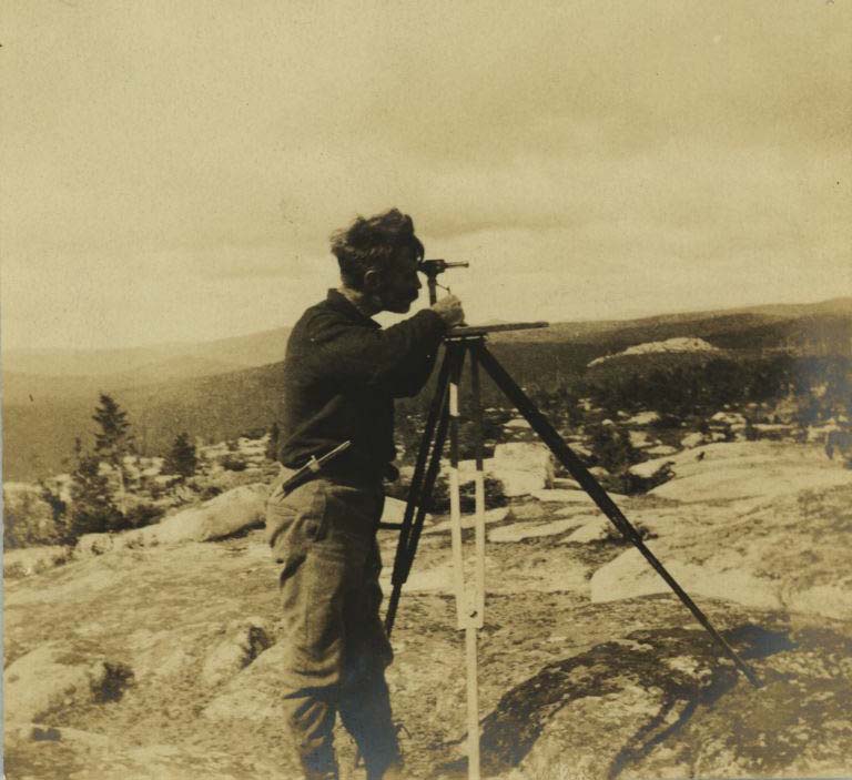 Photographie noir et blanc d’un homme sur des rochers avec de l’équipement d’arpentage